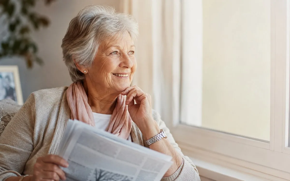 Mujer mayor sonriente leyendo el diario junto a la ventana
